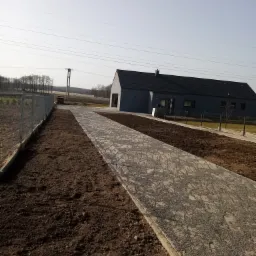 Ułożona ścieżka z kostki brukowej prowadząca do nowoczesnego domu z szarą elewacją, obok świeżo przygotowany teren pod trawnik i ogrodzenie panelowe.