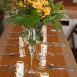 Uroczysta aranżacja stołu z żółtymi kwiatami w szklanym wazonie na wysokiej nóżce, obok zwinięte serwetki z motywem kwiatowym i srebrnymi sztućcami na brązowym bieżniku.