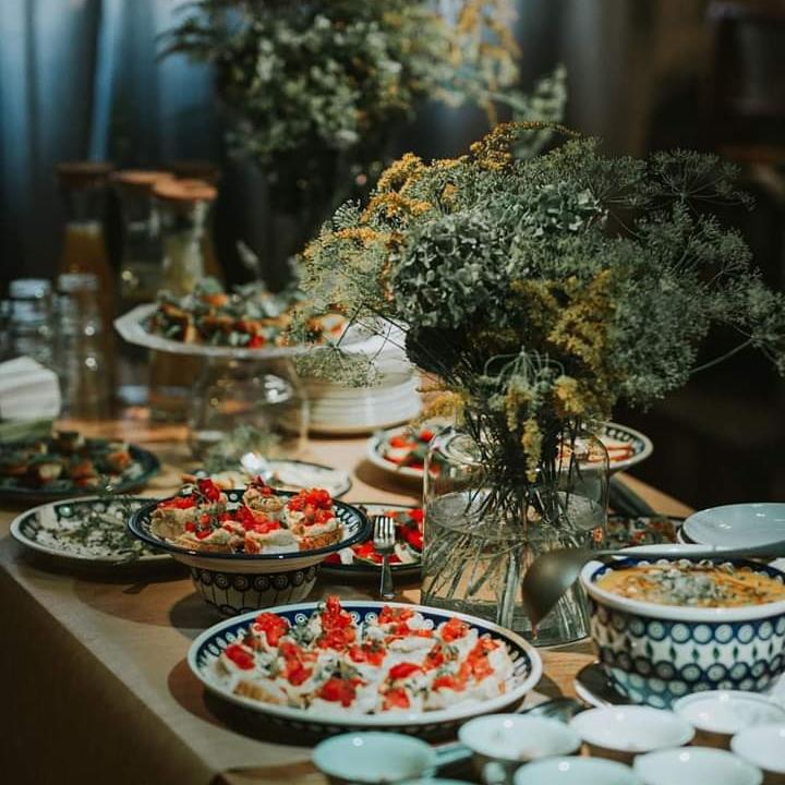 Stół z przekąskami w stylu rustykalnym: talerze z bruschettą, sałatki, zupa w miseczkach z niebieskim wzorem, bukiet polnych kwiatów w szklanym wazonie.