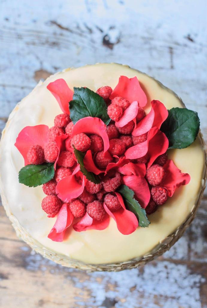 Tort pokryty kremem, udekorowany świeżymi malinami, płatkami róż i zielonymi liśćmi, ujęcie z góry na drewnianym stole.