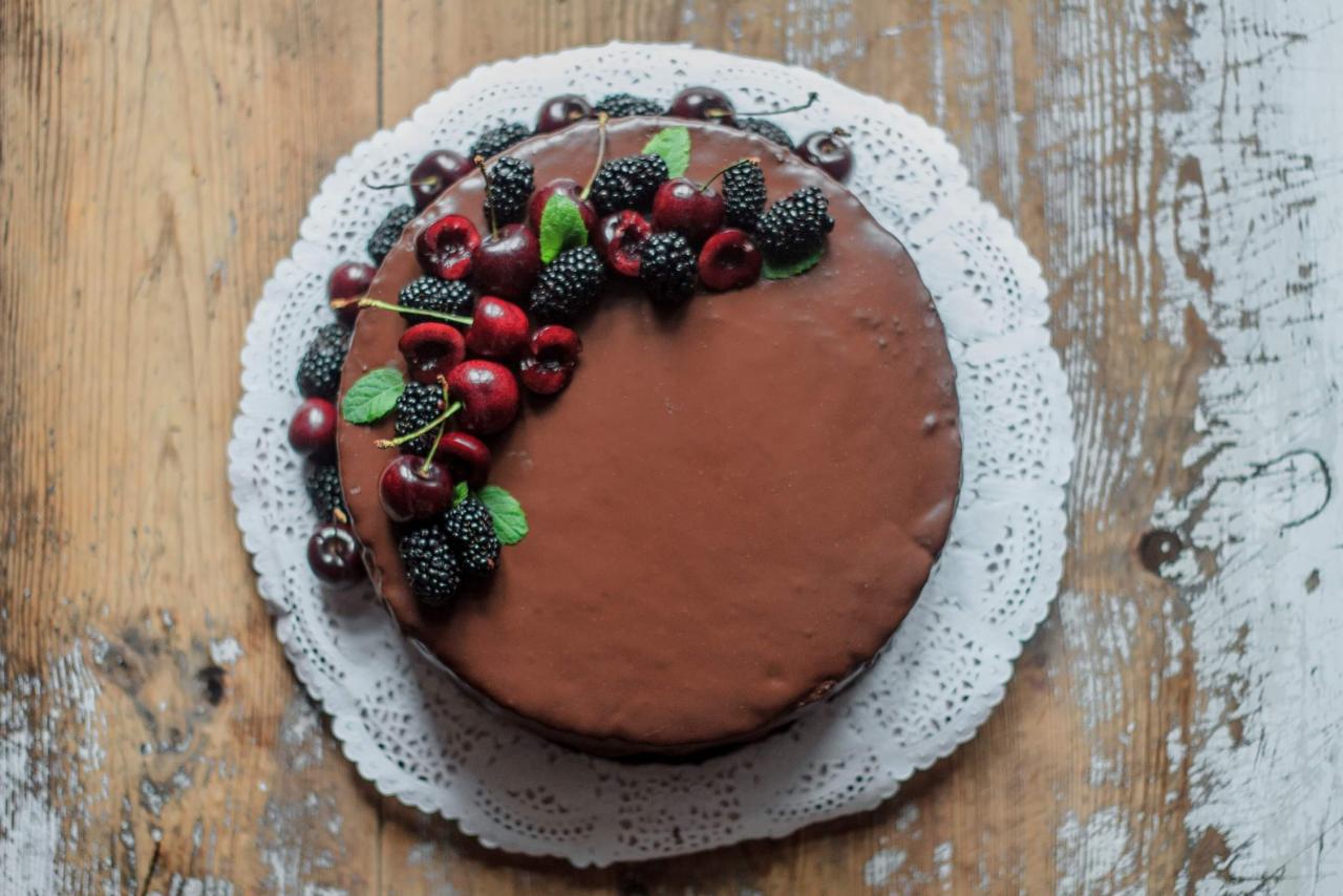Czekoladowy tort udekorowany świeżymi wiśniami i jeżynami, ułożony na białej, ażurowej serwetce na tle drewnianego stołu z widoczną fakturą.