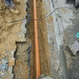 Pomarańczowa rura ułożona w wykopie ziemnym, widoczne fragmenty kostki brukowej i betonowego obrzeża, pracownik w ubraniu moro i czerwonej rękawicy.