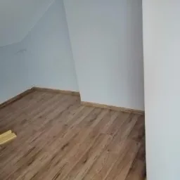 Poddasze po remoncie: świeżo położona podłoga laminowana o wzorze drewna i pomalowane na biało ściany w skosach.