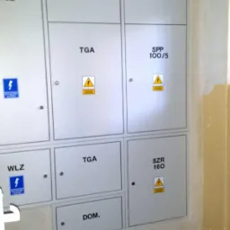 Zestaw skrzynek elektrycznych z oznaczeniami Wlz, Ta, Pomiar, Tga, Spp 100/5, Szr 160 i Dom, umieszczonych na ścianie w korytarzu.