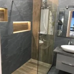 Nowoczesna kabina prysznicowa walk-in z szarymi płytkami imitującymi kamień, drewnopodobną podłogą, oświetlonymi wnękami i szklaną ścianką.