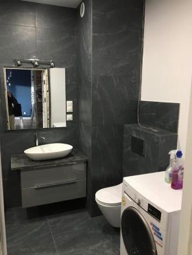 Nowoczesna łazienka z ciemnymi płytkami imitującymi marmur, umywalką nablatową w kształcie misy, szafką pod umywalką z szufladami, lustrem z oświetleniem oraz pralką z detergentami na blacie.