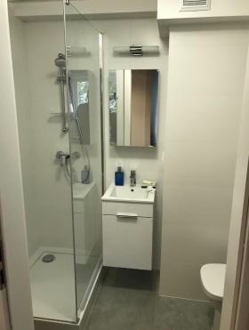 Nowoczesna, jasna łazienka z kabiną prysznicową ze szklaną ścianką, umywalką z szafką w zabudowie i lustrem z oświetleniem. Widoczna minimalistyczna armatura i szare płytki podłogowe.