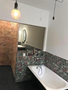 Narożna wanna obudowana płytkami w kwiaty, umywalka z lustrem w tle, fragment ściany z czerwonej cegły, surowa żarówka na kablu.