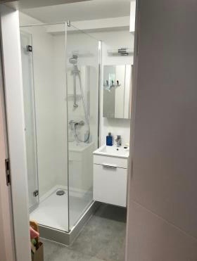 Nowoczesna, mała łazienka z kabiną prysznicową ze szklanymi drzwiami, białą umywalką z szafką i lustrem z oświetleniem. Widok zza uchylonych drzwi.