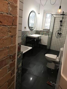 Nowoczesna łazienka po remoncie z elementami cegły, czarną podłogą, owalnym lustrem, kabiną prysznicową typu walk-in z czarną armaturą i wiszącą lampą z żarówką Edisona.