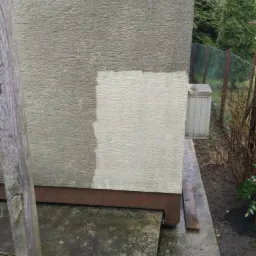 Fragment elewacji budynku z widoczną różnicą przed i po czyszczeniu, z wyraźnie jaśniejszym, umytym kwadratem na szarej, chropowatej powierzchni, z drewnianym słupkiem z lewej strony i zielenią w tle.