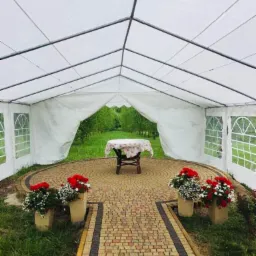 Biały namiot z otwartym tyłem w Lubeni, prowadzący na zewnątrz ścieżką z kostki brukowej, z donicami kwiatów po bokach i stolikiem w tle.