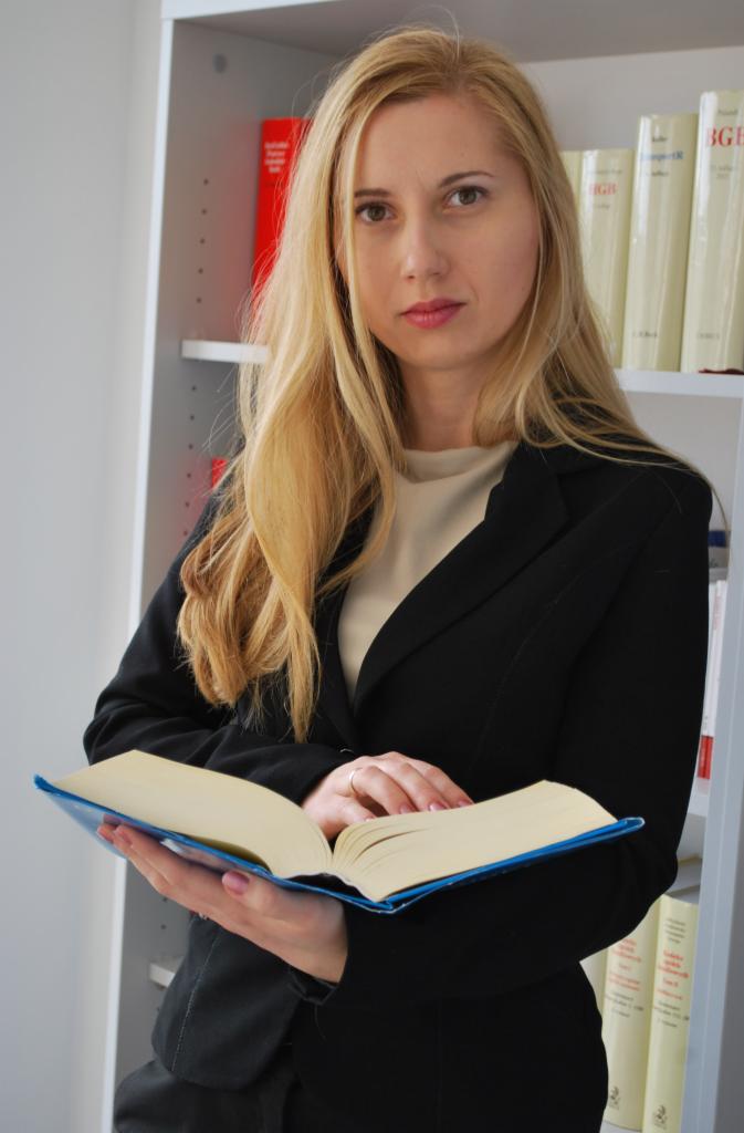 Portret kobiety w czarnym żakiecie, trzymającej otwartą książkę z niebieską okładką na tle regału z książkami w stonowanych kolorach.