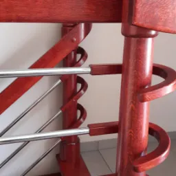 Detal rustykalnej klatki schodowej z drewnianą balustradą w odcieniach czerwieni i metalowymi prętami, widok z bliska na ozdobne elementy i strukturę drewna.