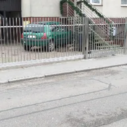 Nowoczesna przesuwna brama posesyjna ze stali nierdzewnej z pionowymi prętami, widok z ulicy na podjazd z zaparkowanymi samochodami i wejście do domu z dekoracją świąteczną na schodach.