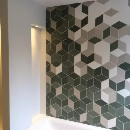 Ściana w łazience po remoncie w Biłgoraju, wyłożona geometrycznymi płytkami w odcieniach szarości, beżu i zieleni, tworzącymi iluzję trójwymiarowych kostek. W tle widoczne oświetlenie niszowe.