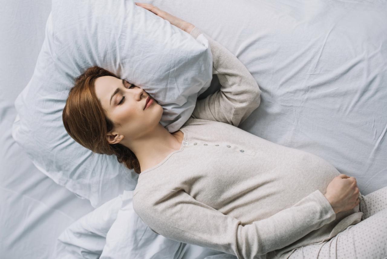 Śpiąca kobieta w ciąży leżąca na białej pościeli i poduszce, ubrana w beżową koszulę nocną z długim rękawem.