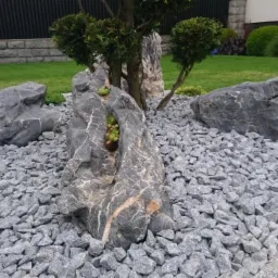 Dekoracyjny klomb z szarych kamieni i dużych głazów, w jednym z nich rosną sukulenty, w tle trawnik i formowane krzewy.