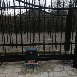 Czarny automat do bramy przesuwnej BFT Ultra zamontowany na betonowej kostce brukowej, widoczne przewody w niebieskich peszlach i elementy mocujące do bramy.
