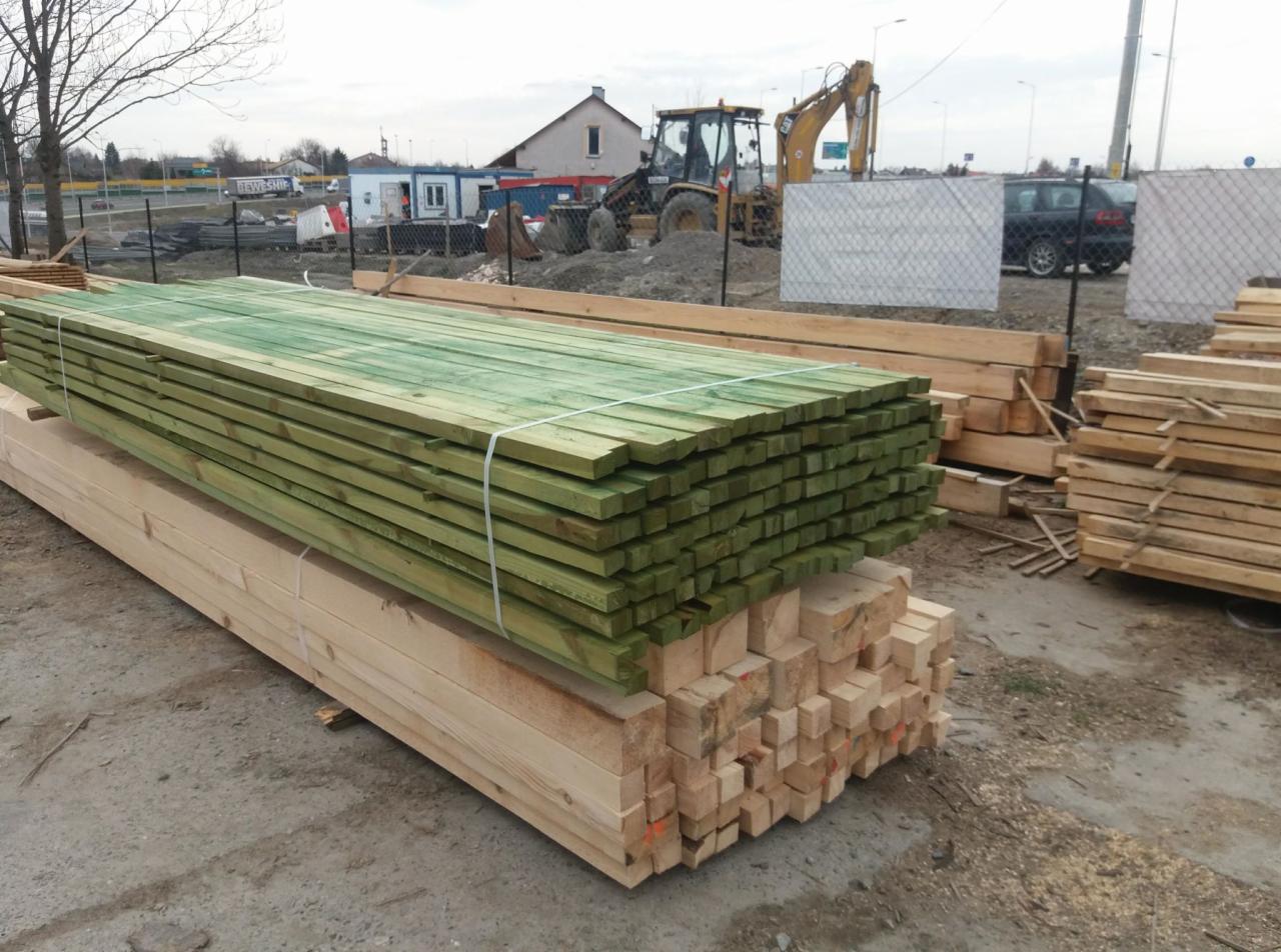 Stosy impregnowanych na zielono desek i kantówek ułożone na placu budowy, w tle widoczny budynek i koparka.