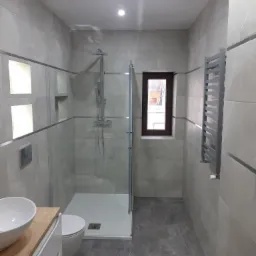 Nowoczesna łazienka z kabiną prysznicową typu walk-in, szarymi płytkami z delikatnym wzorem, grzejnikiem drabinkowym i drewnianym blatem pod umywalką.