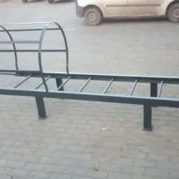 Szara metalowa konstrukcja przypominająca tunel nad przejściem, z elementami drabiny, na tle szarej kostki brukowej i zaparkowanego samochodu dostawczego.