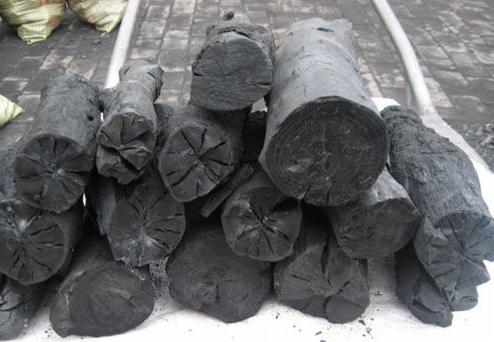 Stos ułożonych kawałków czarnego węgla drzewnego, z widocznymi pęknięciami i strukturą drewna, na tle worków i białej rury.