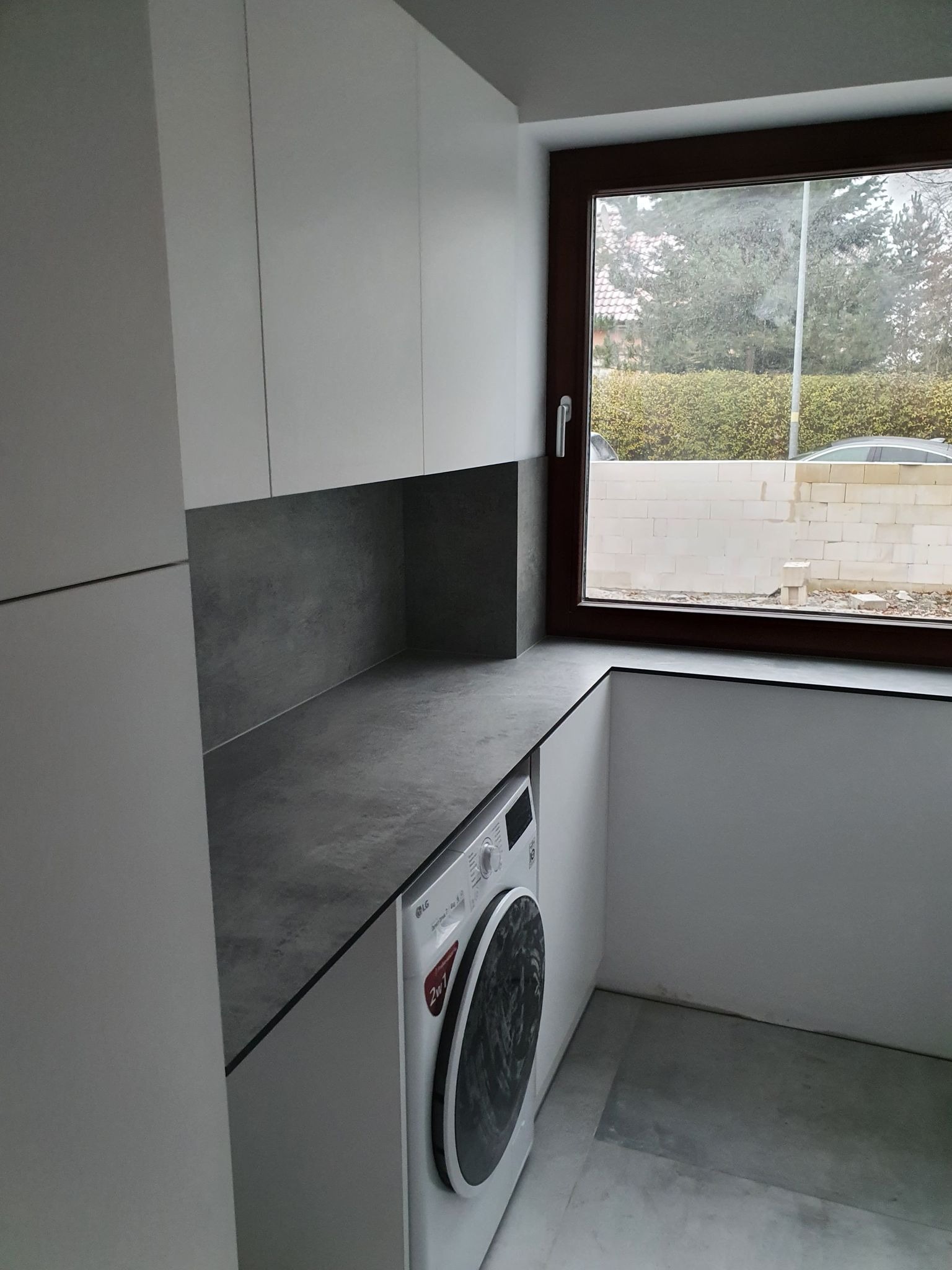 Nowoczesna aranżacja pralni w Ośnie Lubuskim: białe szafki wiszące, szary blat, pralka LG pod blatem, okno z widokiem na ogród w trakcie budowy.