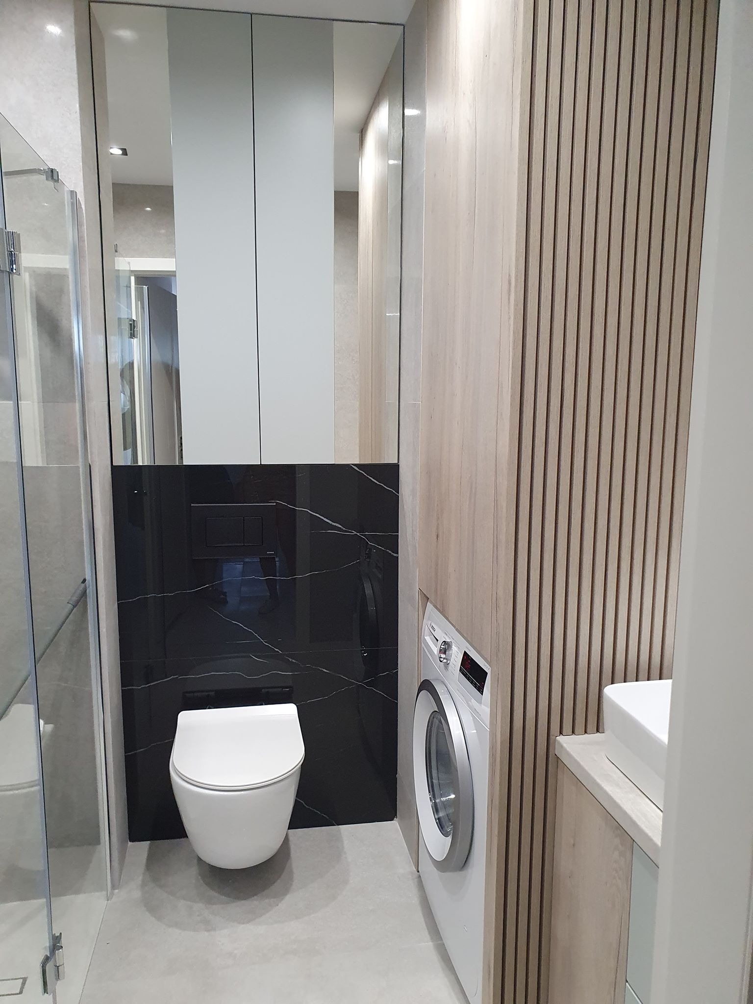 Nowoczesna łazienka z toaletą podwieszaną na tle czarnej płyty z białym żyłkowaniem, pralka obok drewnianej ściany z pionowych paneli, widoczna szklana kabina prysznicowa.
