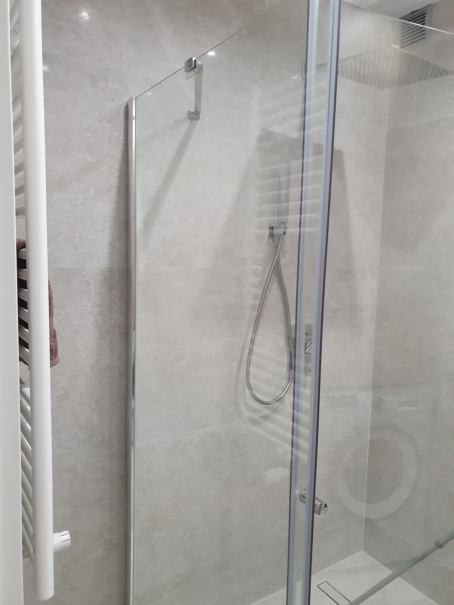 Nowoczesna kabina prysznicowa z deszczownicą, szklane drzwi z chromowanymi zawiasami, grzejnik drabinkowy z ręcznikiem w tle, szare płytki imitujące kamień na ścianach.