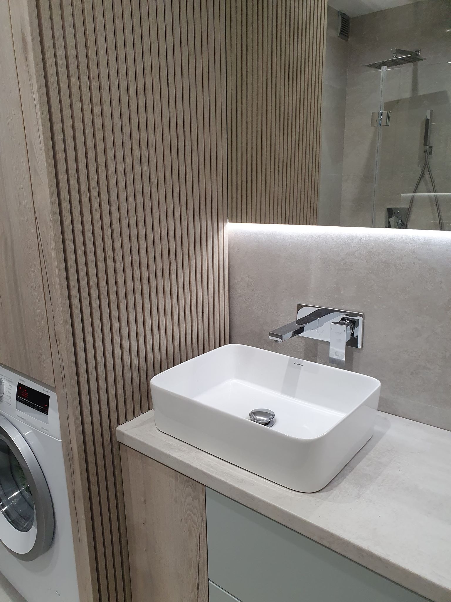 Nowoczesna łazienka z umywalką nablatową o kwadratowym kształcie, chromowaną baterią ścienną, drewnianą ścianą z pionowych listew i pralką w tle.