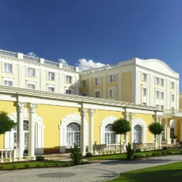 Elegancki, żółto-biały budynek hotelowy z kolumnami i balkonami, otoczony zielonym trawnikiem i drzewami w słoneczny dzień.