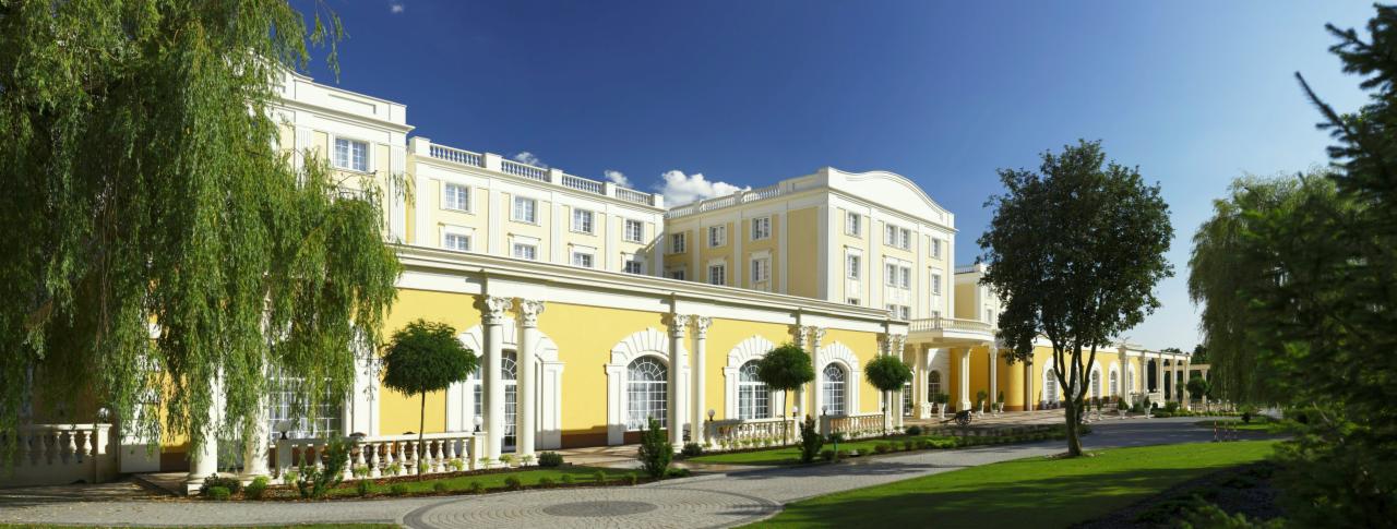 Elegancki, żółto-biały budynek hotelowy z kolumnami i balkonami, otoczony zielonym trawnikiem i drzewami w słoneczny dzień.