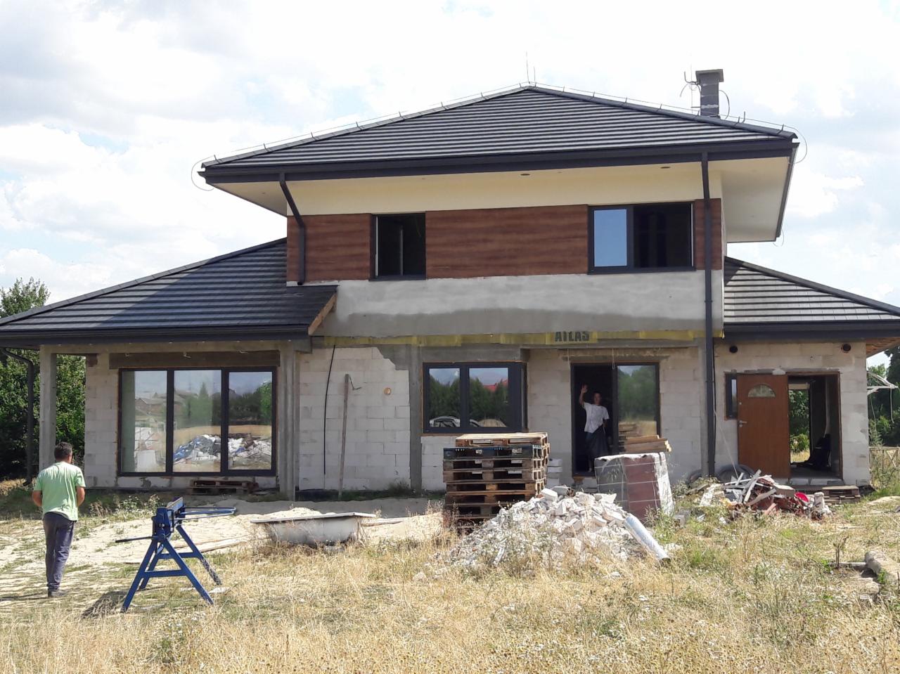 Dom jednorodzinny w trakcie budowy z ciemną dachówką, widoczne palety, gruz i pracownik machający w drzwiach.