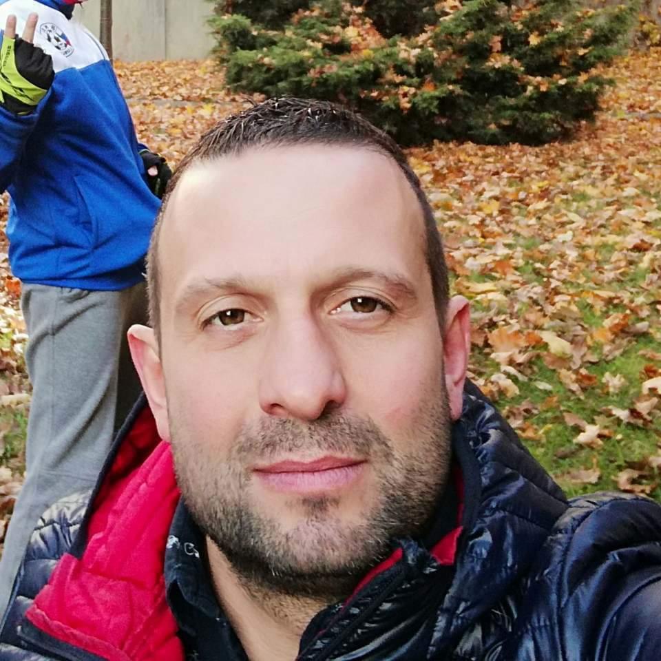 Portret mężczyzny w kurtce na tle opadłych liści i krzewów, z drugą osobą częściowo widoczną w tle.