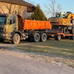 FHU ROMAN LACH - Ciężarówka Scania z pomarańczową skrzynią ładunkową przewozi na niskopodwoziowej przyczepie żółtą koparkę Liebherr 904 na tle domu i drzew.