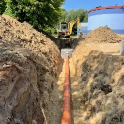 Długi, pomarańczowy rurociąg ułożony w głębokim wykopie ziemnym, z widoczną koparką w tle i betonowym kręgiem studziennym. Na drugim planie zbiornik na wodę.