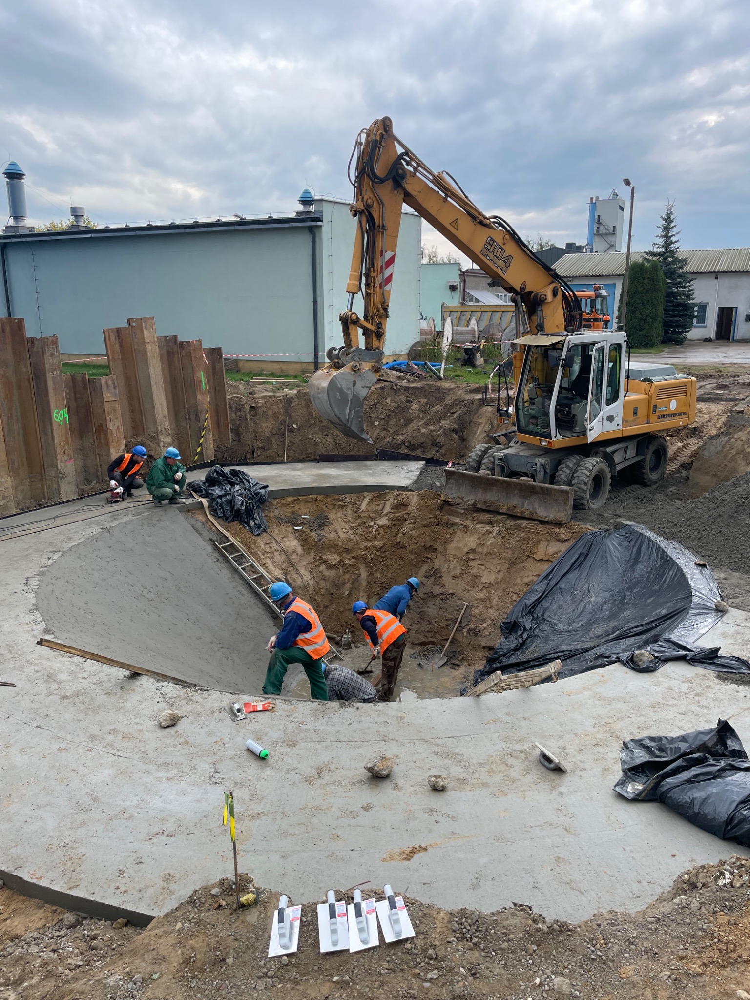 Prace ziemne przy okrągłym wykopie zabezpieczonym ściankami Larsena, na dnie wylany beton, robotnicy w kaskach i kamizelkach odblaskowych, koparka Liebherr 904 w tle, pochmurne niebo.