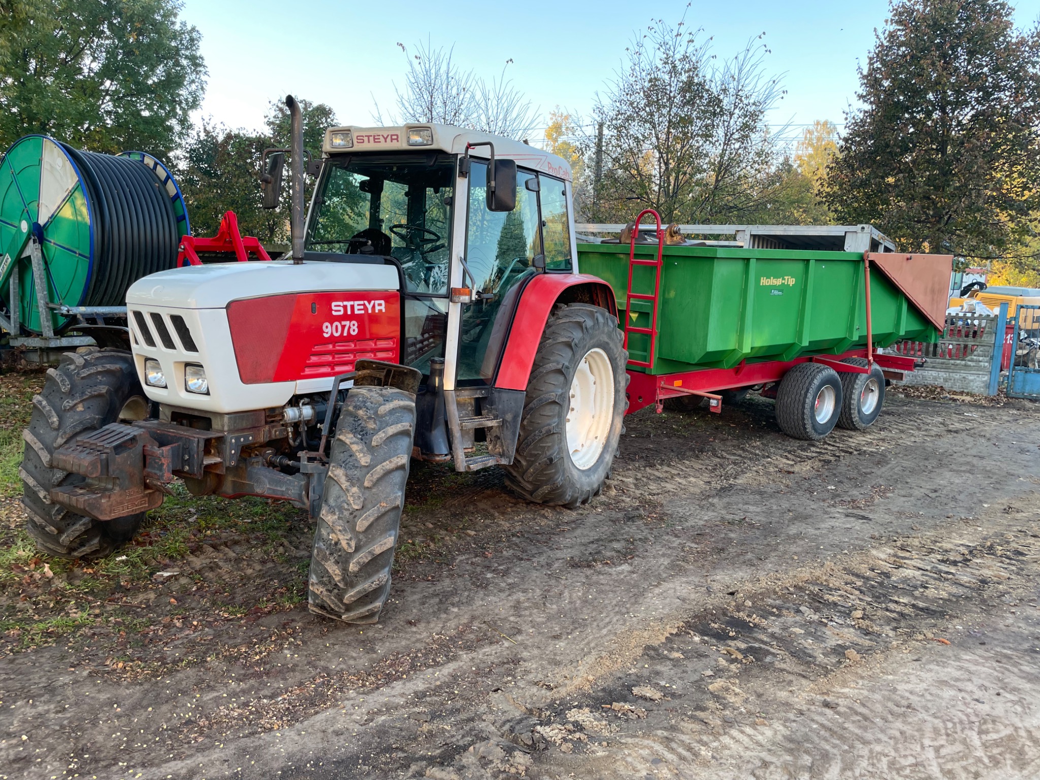Czerwono-biały traktor Steyr 9078 z zieloną przyczepą Holsa-Tip na błotnistym terenie, z rolką kabla w tle.