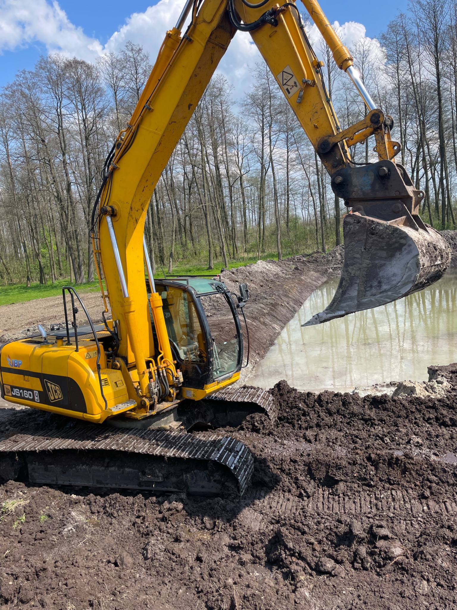 Żółta koparka JCB JS160 głębi staw, widoczne ślady gąsienic na błotnistym brzegu i drzewa w tle.