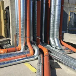 Instalacja rur wentylacyjnych z tworzywa sztucznego i metalu w budynku w stanie surowym, widoczne izolacje ścian z wełny mineralnej i fragment pracownika w niebieskim kombinezonie.