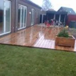 Błyszczący, drewniany taras przydomowy z donicą z małym drzewkiem, widoczny trawnik i budynek z dużymi oknami przesuwnymi.