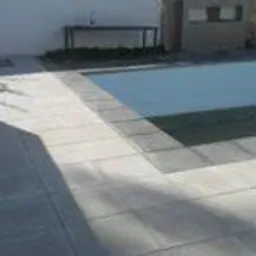 Jasnoszara kostka brukowa ułożona wokół basenu, widoczny fragment białego muru i zieleń w tle.