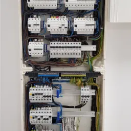 Rozdzielnica elektryczna w białej obudowie, widoczne liczne bezpieczniki i okablowanie, niekt&oacute;re z diodami LED sygnalizującymi stan, zamontowana na białej ścianie, z widocznym logo firmy w prawym...