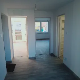 Wnętrze mieszkania po odświeżeniu: białe ściany, szara podłoga imitująca drewno, widok na balkon i inne pomieszczenia przez otwarte drzwi.