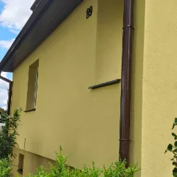 Żółta elewacja domu z brązowymi rynnami i numerem 89, widoczna z poziomu krzewów. Ujęcie z boku, w tle fragment nieba z chmurami.