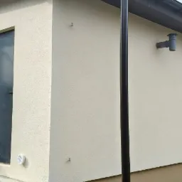 Elewacja budynku po ociepleniu i pomalowaniu na beżowo. Widoczny fragment dachu, rynna, pionowa rura spustowa, drzwi wejściowe oraz fragment samochodu dostawczego.