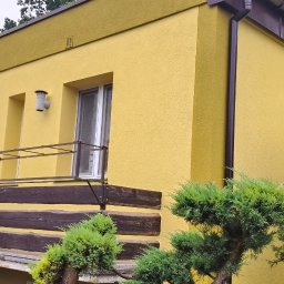 ADEXBUD Usługi Remontowo-Budowlane - Żółty dom z balkonem i brązowymi elementami drewnianymi oraz brązową rynną. W tle zieleń drzew. Na dachu antena.