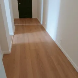 Jasne panele podłogowe w korytarzu z ciemnymi drzwiami. Nowoczesne wykończenie wnętrza z widocznymi listwami przypodłogowymi i gładkimi, białymi ścianami.