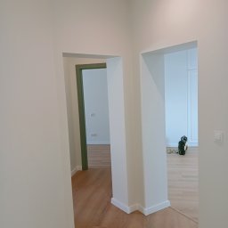 ADEXBUD Usługi Remontowo-Budowlane - Białe wnętrze po remoncie: świeżo pomalowane ściany, drewnopodobna podłoga. W tle widoczne zielone drzwi i narzędzia malarskie.